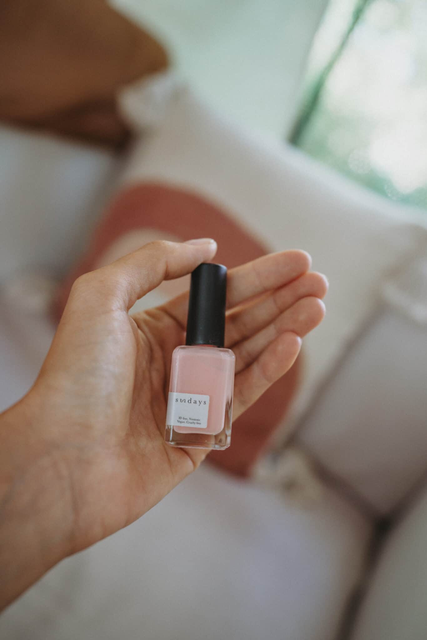 A hand showcasing a plastic-free pink nail polish bottle in front of a couch.