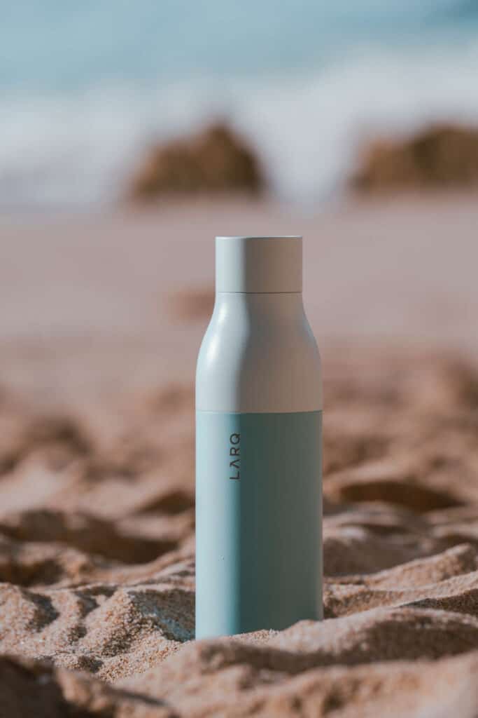 An eco traveler's blue bottle resting in the sand near the ocean.