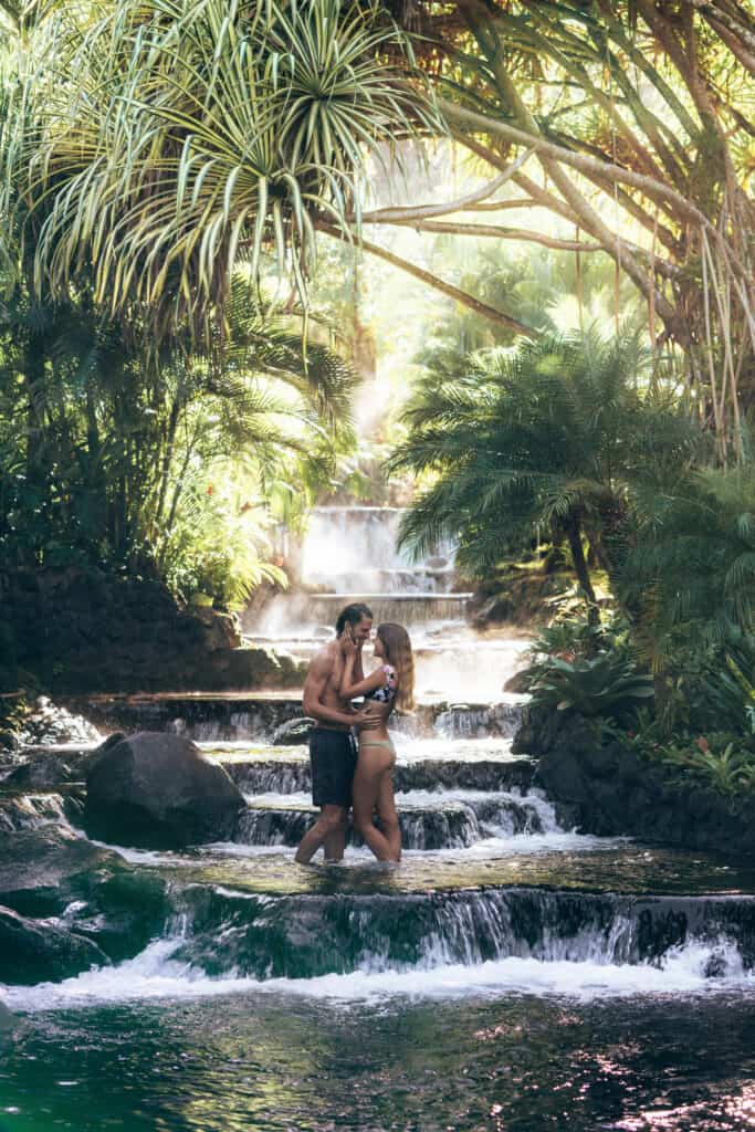 A couple kisses in front of a waterfall in the jungle.