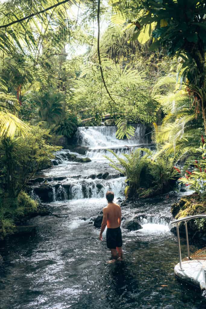 A man wading in a stream near a waterfall.