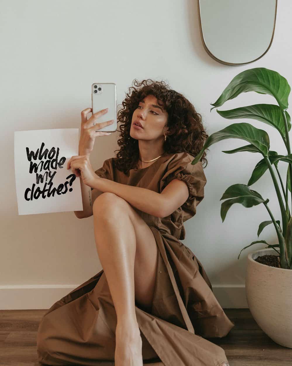 A sustainability advocate sitting on the floor holding a sign questioning the necessity of clothing.