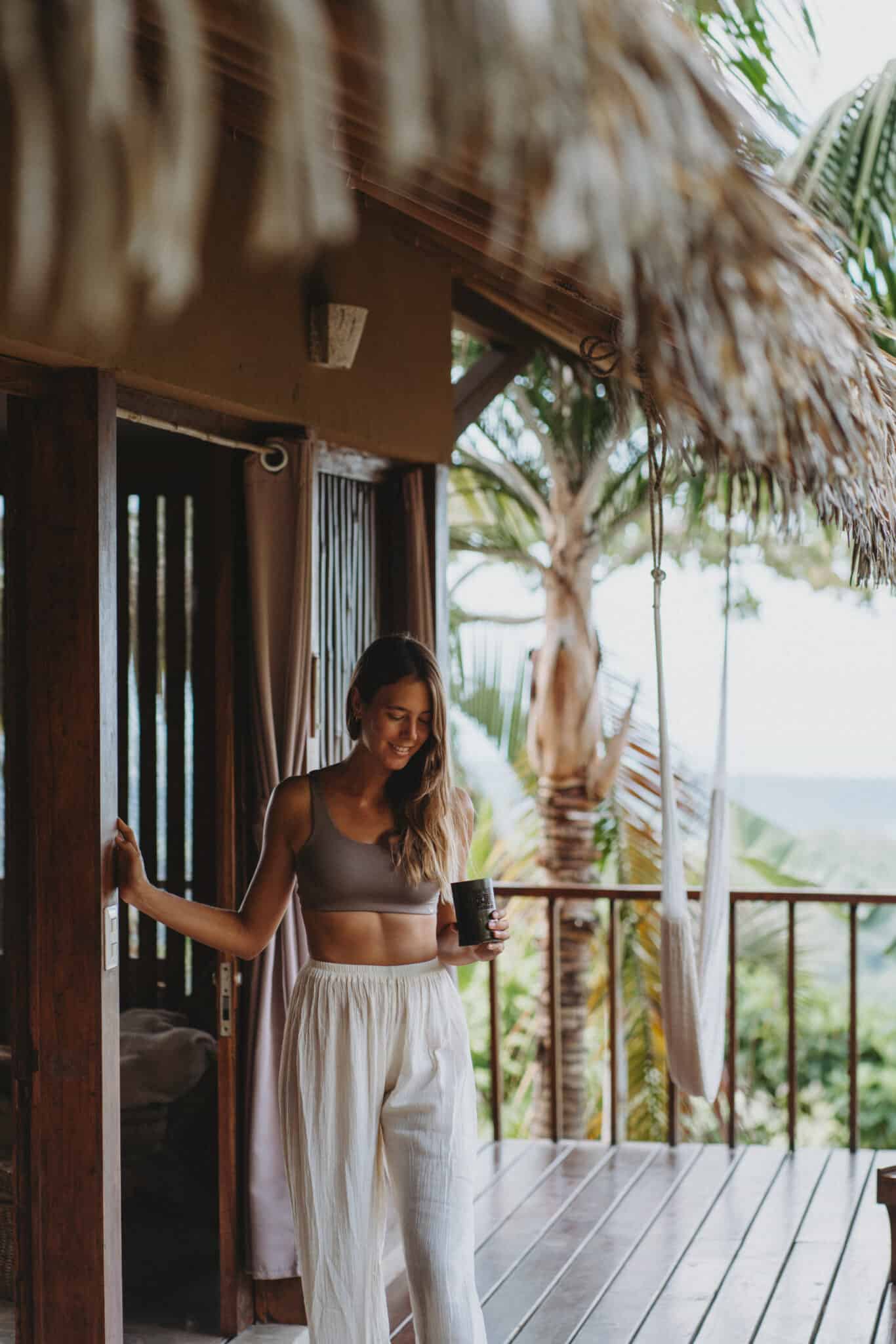 A woman, standing on the porch of a house, invites you to work with us amidst the thatched roof.