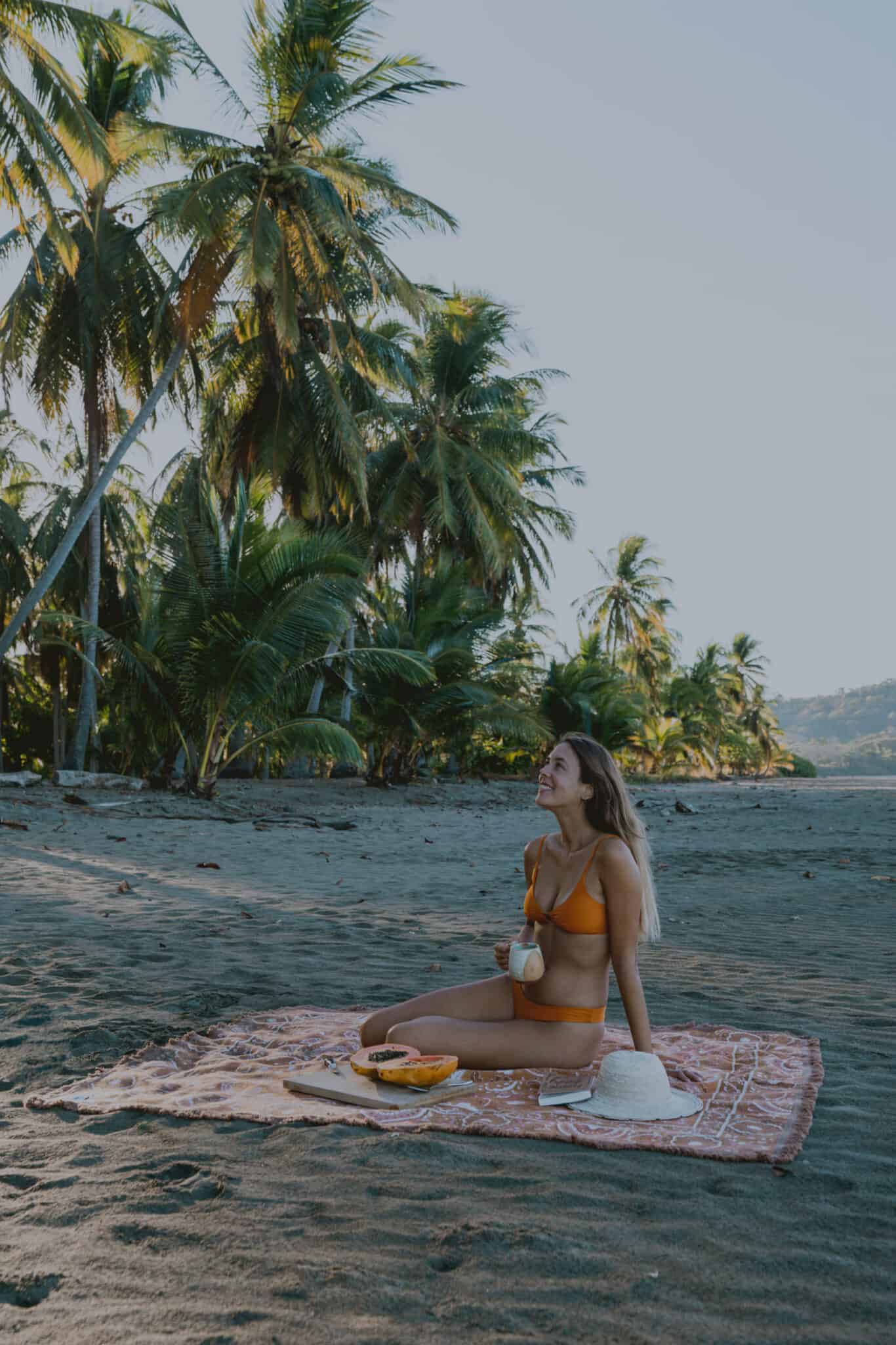 A woman in a bikini sits on a blanket at the beach, inviting you to work with us.