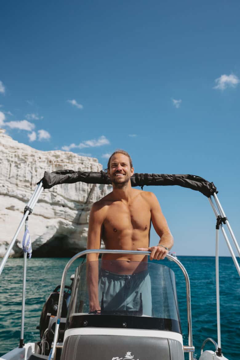 A shirtless man standing on the back of a boat in Milos, Greece.