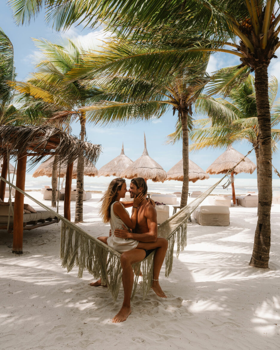 A couple sitting in a hammock on the beach under palm trees.