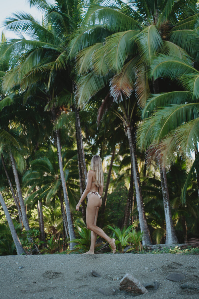 A woman in a bikini walking on a beach in Santa Teresa, Costa Rica in front of palm trees.